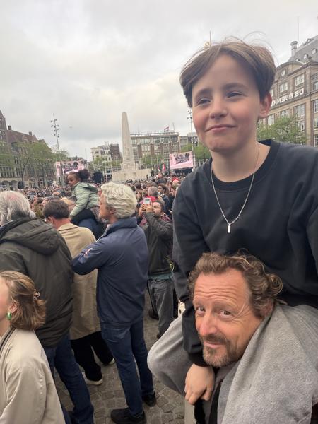 A child sits on a man's shoulders, smiling at the camera amidst a large crowd gathered in Dam Square, Centrum, Amsterdam